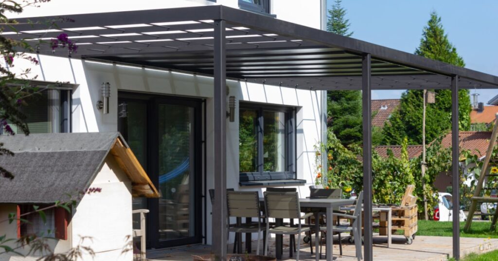 pergola en aluminium noire adossée à une maison blanche avec du mobilier de jardin dessous