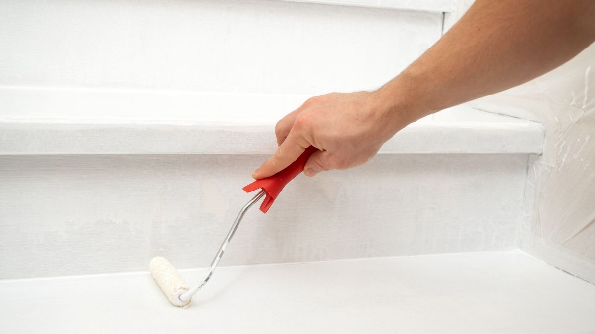 Gros plan sur un bras qui tient un rouleau de peinture et qui peint une marche d'escalier blanc. 