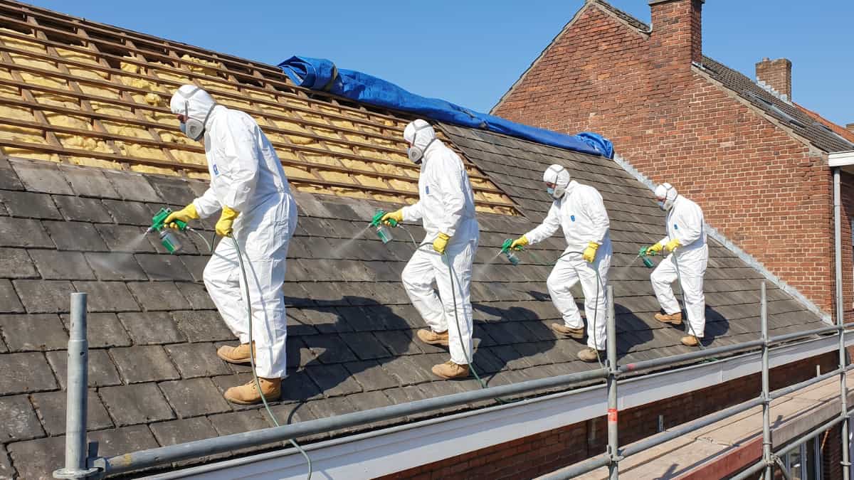 Cinq ouvriers en combinaison blanche, masque respiratoire et gants jaunes se tiennent en ligne sur un toit en pente et pulvérisent un fixateur sur des ardoises en amiante gris foncé à l'aide de pistolets de pulvérisation verts, avec la partie supérieure du toit déjà démontée révélant les lattes et l'isolation jaune sous une bâche bleue, un échafaudage métallique en bas et un mur en briques rouges d'une maison voisine en arrière-plan sous un ciel bleu dégagé.