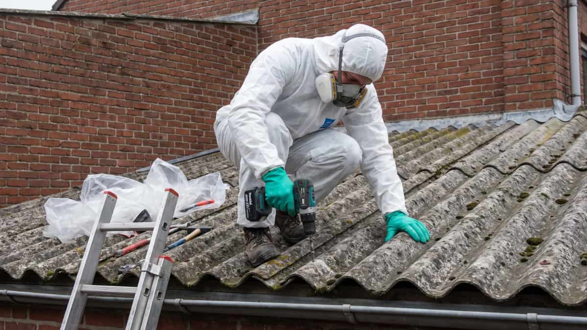 Un ouvrier en combinaison blanche, masque respiratoire et gants verts s'accroupit sur un toit en plaques ondulées de fibrociment recouvertes de mousse et utilise une visseuse sans fil pour démonter une fixation, avec une échelle en aluminium et du film plastique posés à côté de lui devant un mur en briques rouges.